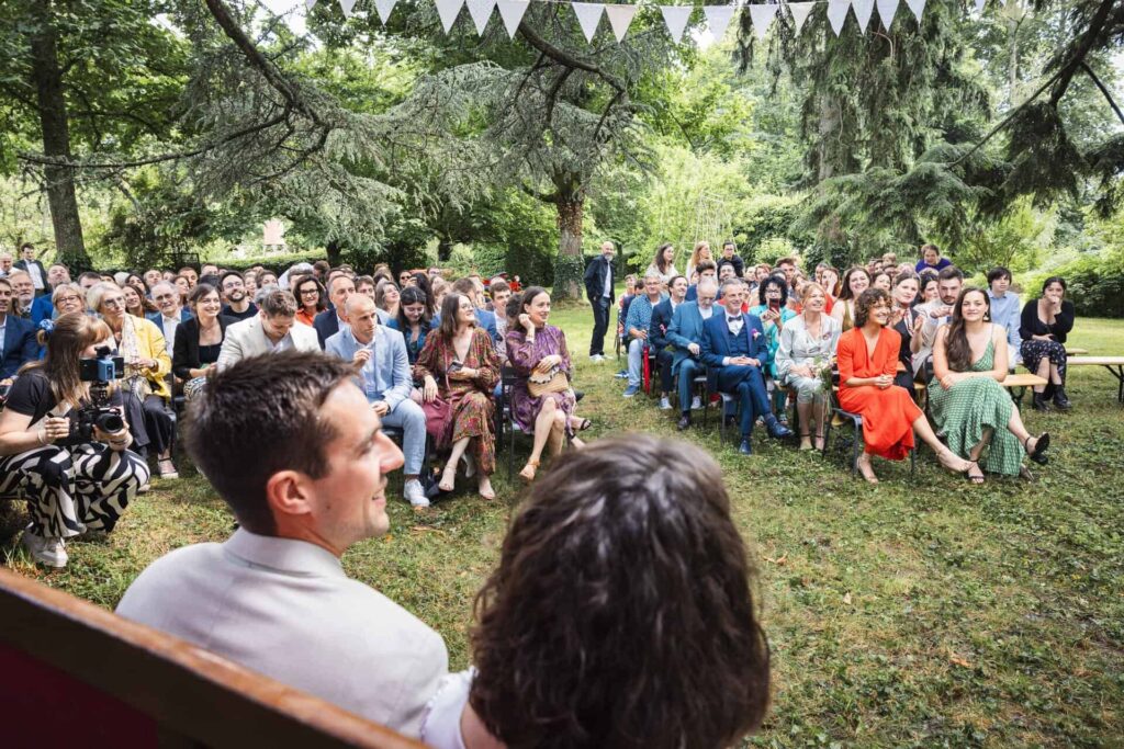 Cérémonie laïque en plein air : les mariés face à leurs invités assis dans l'herbe sous les arbres