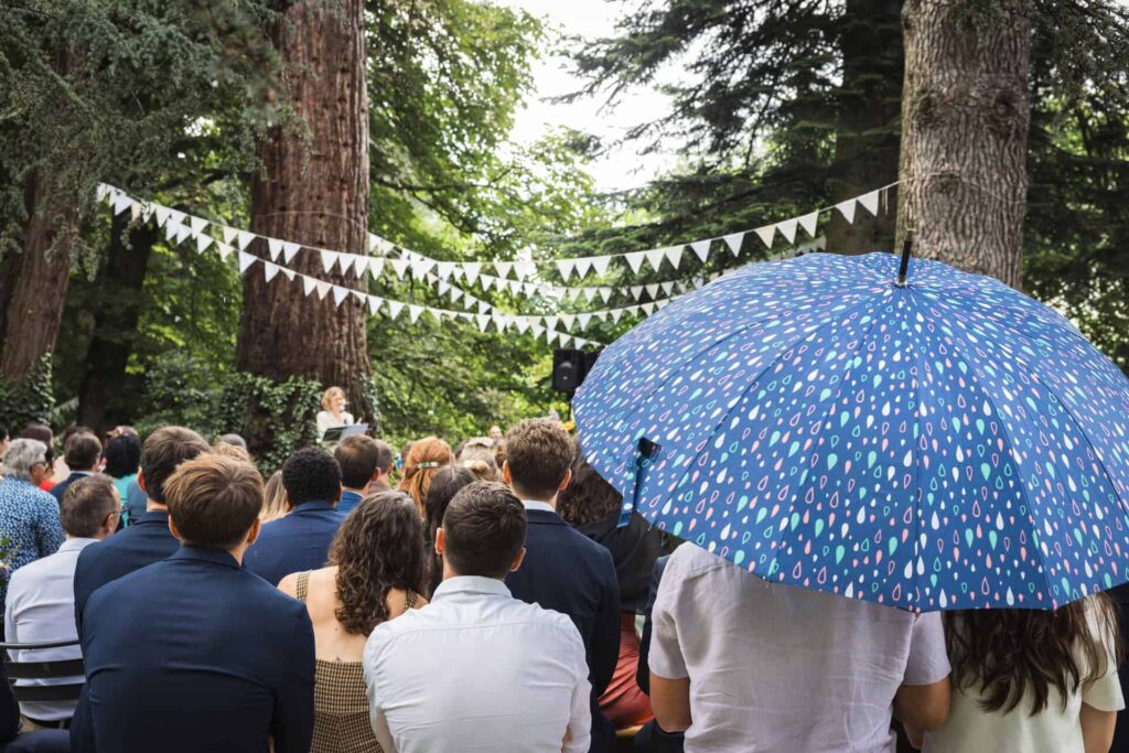 Invités de dos pendant une cérémonie laïque sous les arbres, un parapluie bleu à pois au premier plan, l'officiante au pupitre
